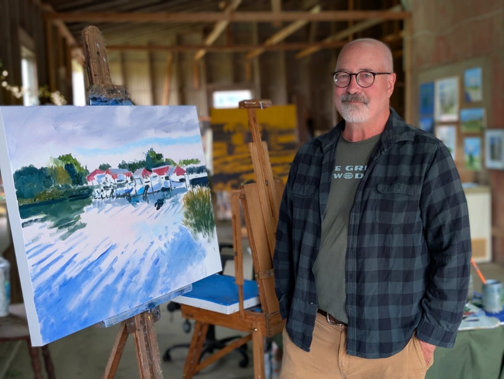 Artist Alan Bull Standing next to his painting of a section of houses on the water